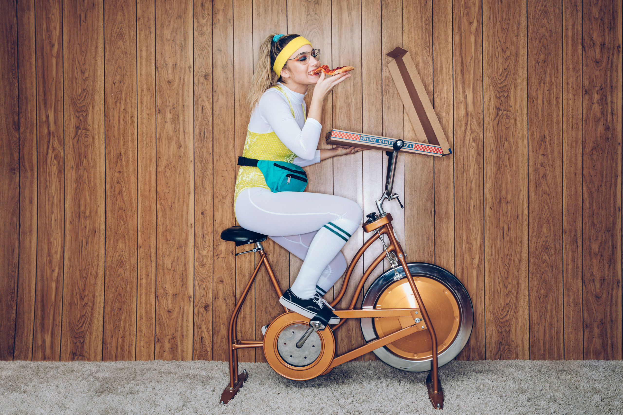 Eine Frau in Trainingskleidung im Stil der 1980er und 1990er Jahre strampelt auf einem stationären Fitnessrad in einem Vintage-Raum mit Zottelteppich und Holzvertäfelung an den Wänden. Sie trägt einen Trikotanzug und eine Gürteltasche und isst aus einem großen Pizzakarton.